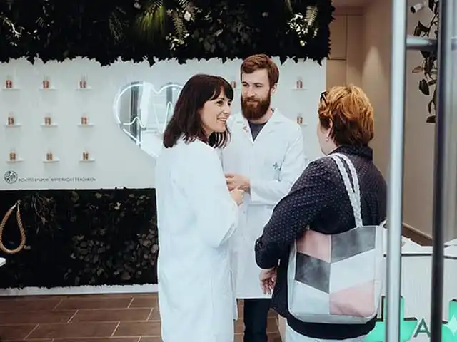 Apothekenteam im Gespräch mit Kundin in moderner Apotheke vor beleuchtetem Herzsymbol